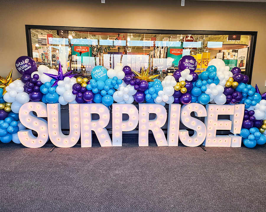 A lighted metal letter sign reading Surprise surrounded by balloons.