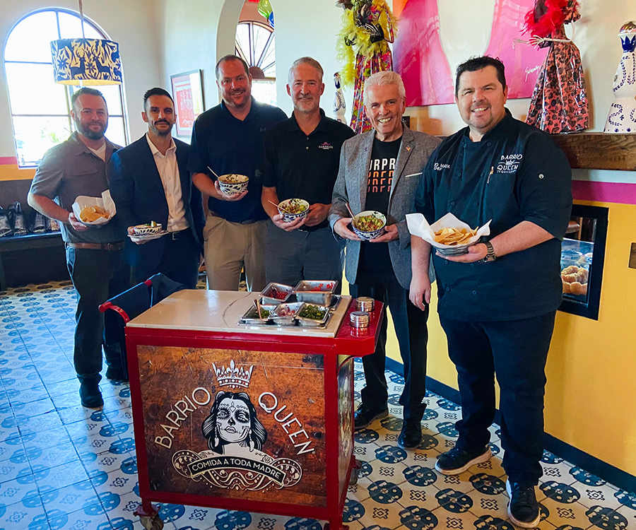 A group of people including Mayor Hall, Vice Mayor Judd and Councilmember Duffy hold bowls of food and pose for a photo around the host station at Barrio Queen in Surprise 
